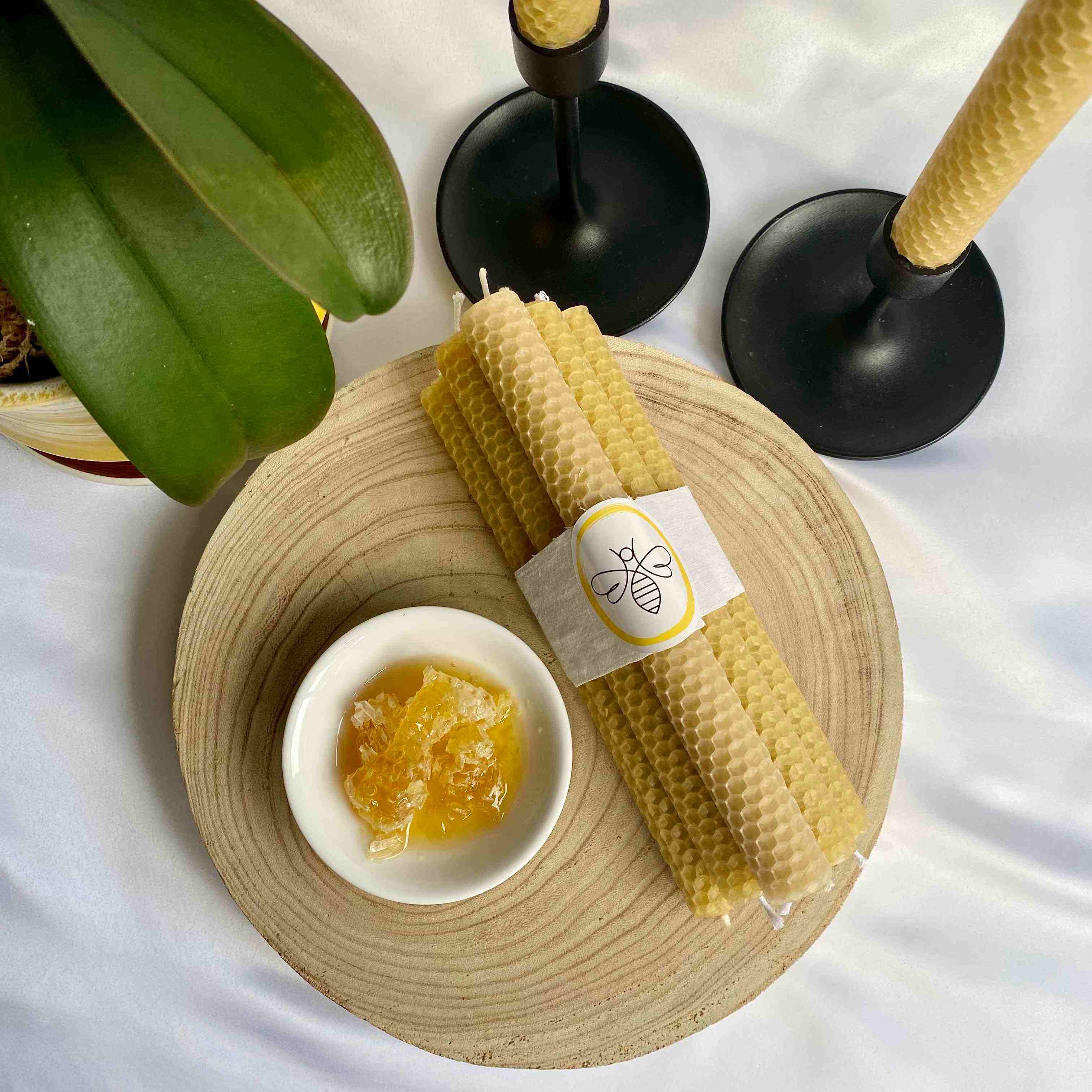 a set of 8 beeswax dinner candles wrapped in white line, next to a bowl of honeycomb two candles in black candle sticksn